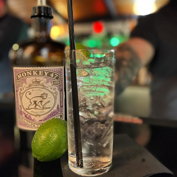A Monkey 47 gin bottle, lime, and a gin and tonic with a straw on a bar.