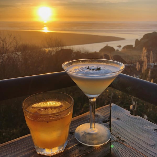 Two cocktails on a wooden railing overlooking a sunset over the ocean and coastline.